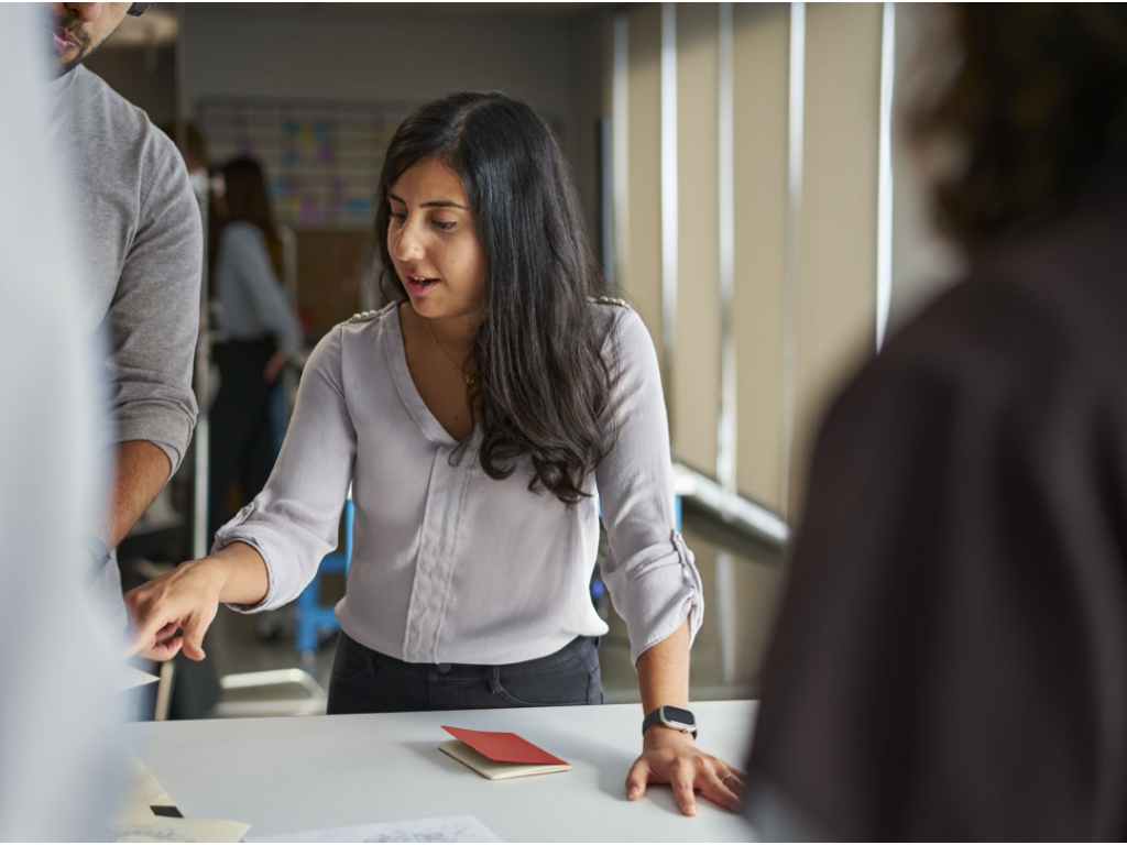 Young woman stands at a table surrounded by people leading a discussion