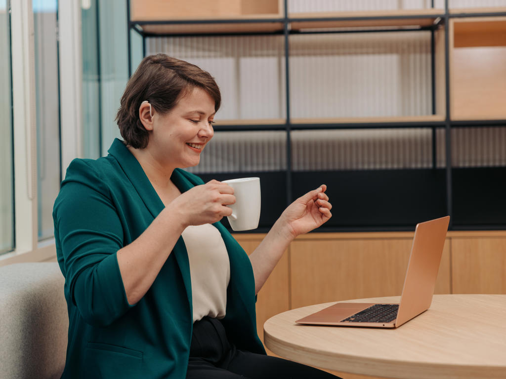 A woman works on her laptop while holding a cup of coffee in an office. Her hair is tucked behind her ear to show off their hearing aid.